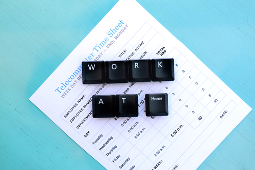 Black keyboard keys spelling "WORK AT Home" on top of a telecommuter timesheet placed on a light blue wooden surface. 