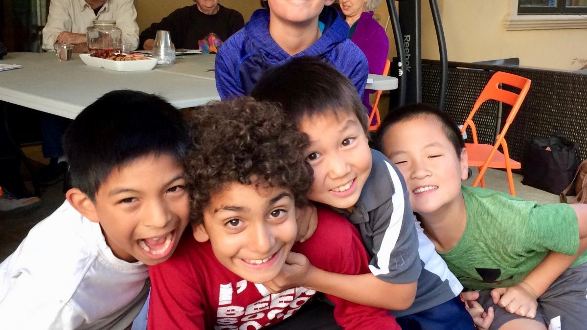 Four young boys huddled together, smiling at the camera during an indoor social gathering.